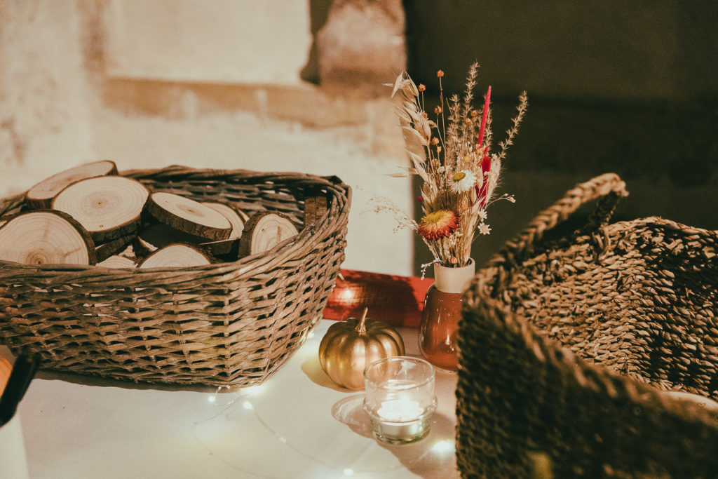 Un décoration de mariage écologique : des paniers en osier de location avec des rondelles de bois, un vase rempli de fleurs séchées, une bougie et guirlande lumineuse sur une table.