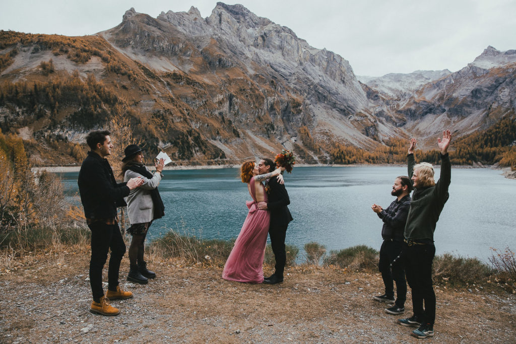 Une des définitions de l'elopement : un elopement au bord d'un lac de montagne, le couple de mariés s'embrasse pendant que 4 invités les acclament.