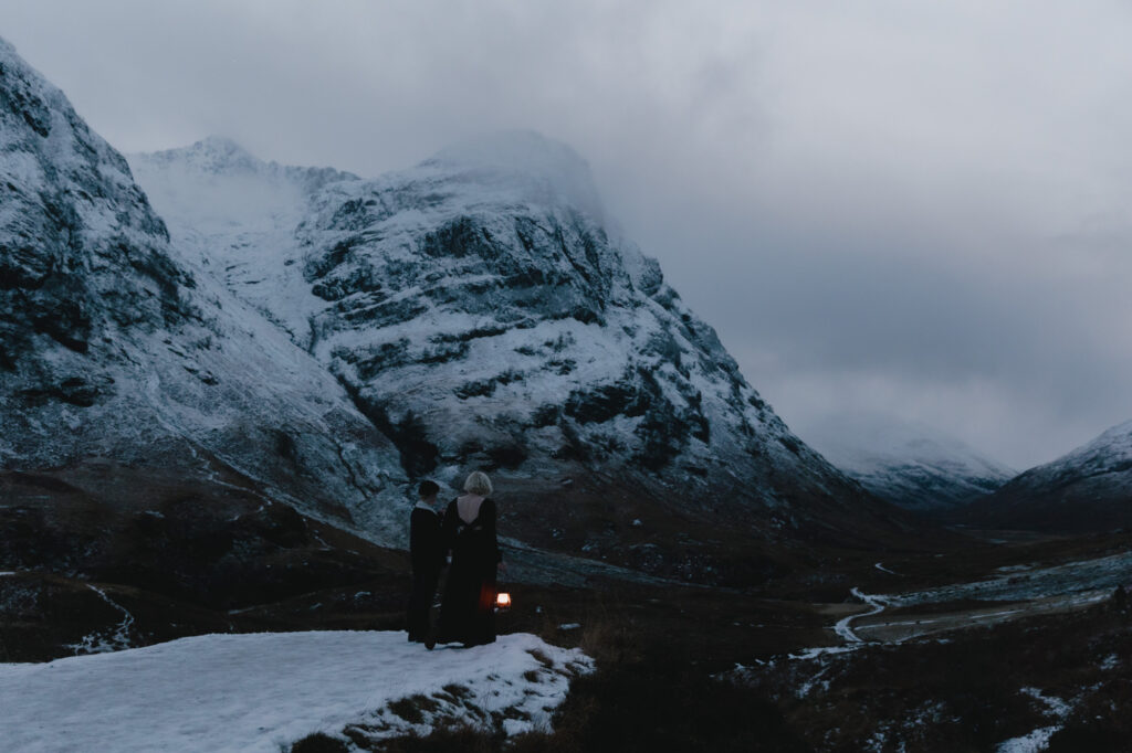 Couple overlooking the valley of Glencoe, with snow-capped mountains in the background. They're holding a lantern.
