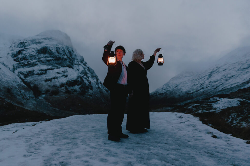 Couple holding lanterns while looking around them, in a snowy landscape in Glencoe, Scotland.