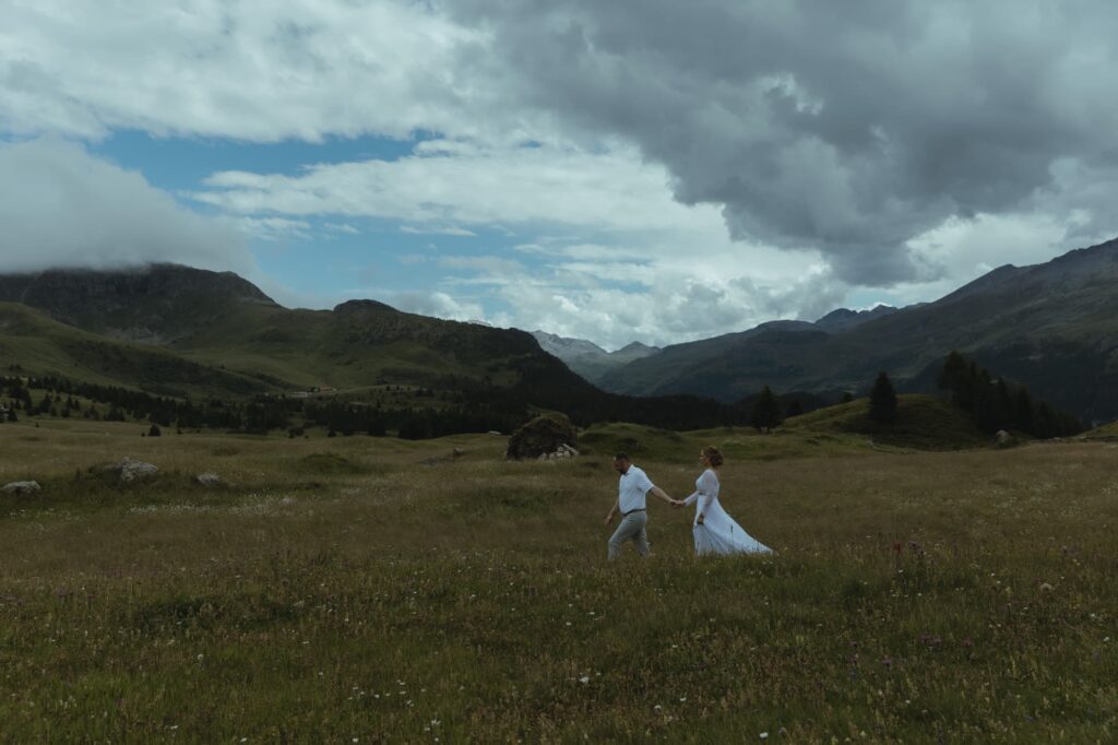 couple holding hands and walking one in front of the other, in a field with switzerland mountains in the background