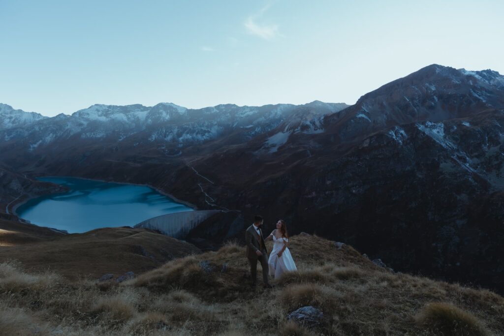 couple hiking up a mountain with a dam an a mountain lake behind them.