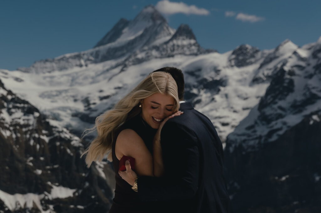 A blonde woman smiling while hugging her partner after he proposed to her, in front of snowy mountains.